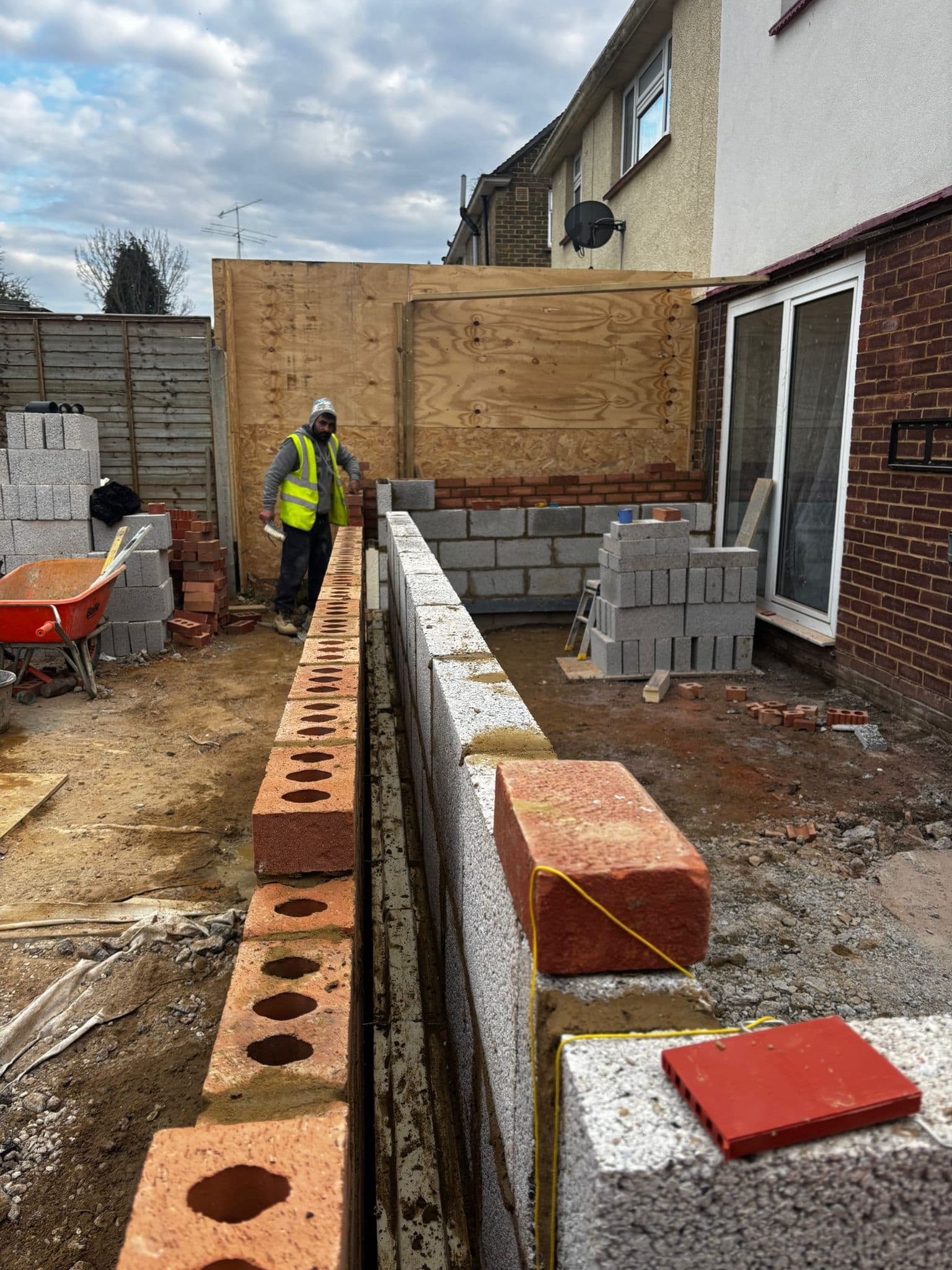 Builder in hi-vis laying blockwork on extension foundations with plywood temporary bracing